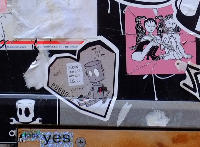 paste up graffiti, one is Robbo-T sitting on ground with his heart in his hand, the other is two young women, one dress in black boots and black outfit, the other is wearing nothing,  Sitting with their knees bent in front of them. 