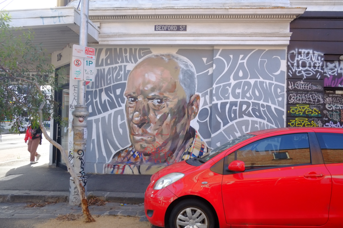 street art painting, portrait by artist LING, of Alf Stewart, under a street sign that says Bedford street, red car parked in front, person on sidewalk beside, 