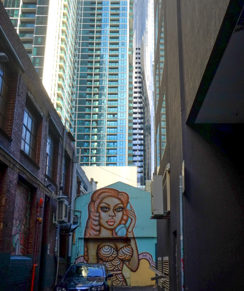 mural of a woman with long orange hair and a tight fitting dress, on a wall at the end of an alley, large condos and apartment buildings rise up behind her
