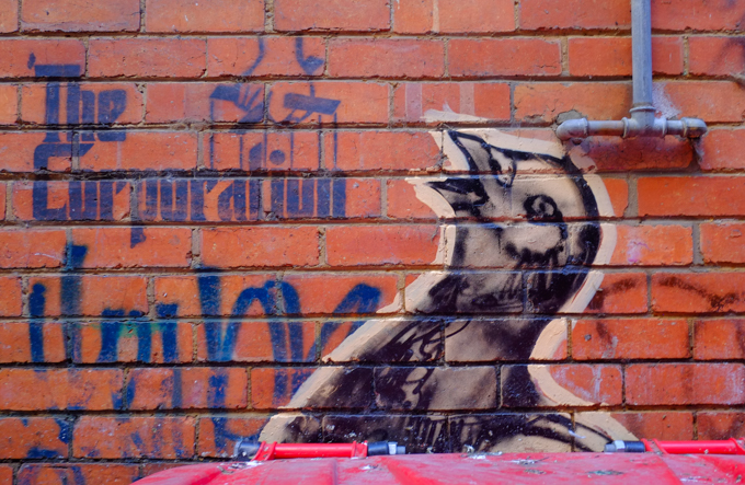 mural, urban art, small black bird looking upwards, with beak open, stencil on brick wall behind says The Corporation, with marionette string