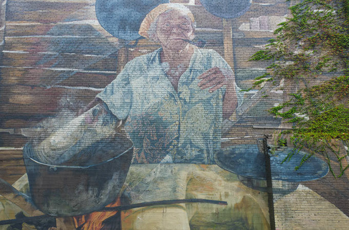 woman in a mural, beside a large table with a black pot on it, head scarf on her head, smoking  a pipe