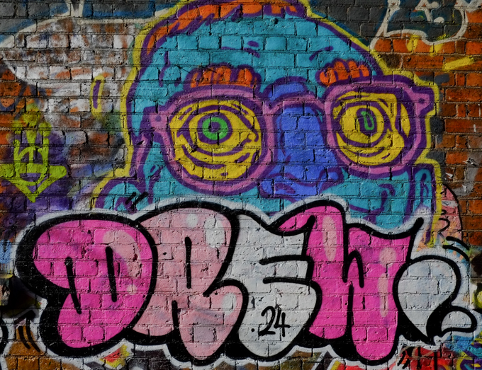 top of head in blue, bright blue nose, pink glasses, yellow and green eyes, red very short hair and red eyebrows, word Drew written across bottom of face, 24