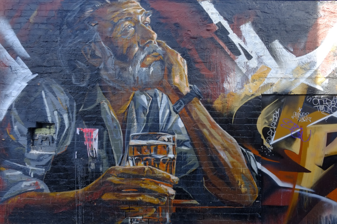 street art painting, portrait by artist LING, of Patrick Coyle, man sitting with a full  glass of beer in his hand, elbow on table, chin in hand, face has mustache and beard