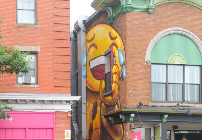 large mural by Spok Brillor of a yellow laughing emoji with tears in one eye, holding a smart phone in his hand.  phone corresponds to a window in the building. 