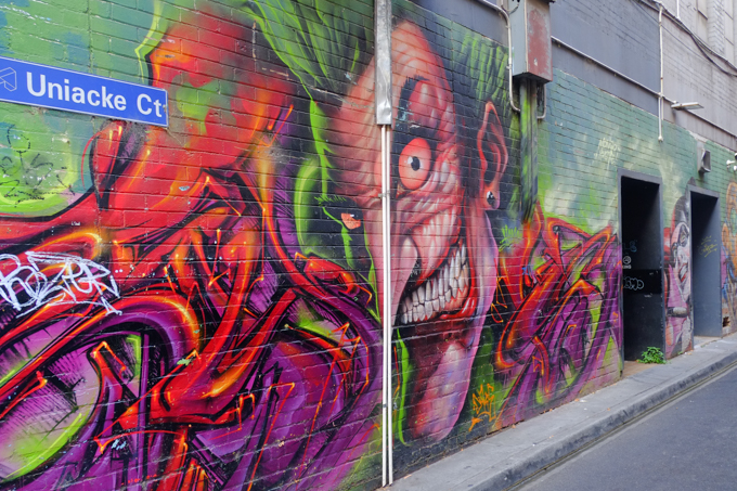 entrance to Uniacke Court, a lane in Melbourne, with a large mural on the wall, ugly man with bulging pink eyes and large teeth, big pointy chin, wild green hair
