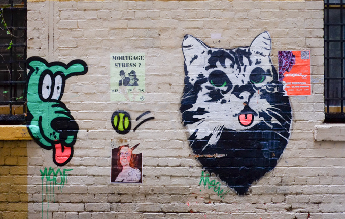 street art on a white painted brick wall, Austral Lane in Melbourne, a green dog head with red tongue hanging out, a black and white cat with a pink tongue, a yellow tennis ball bounces between them
