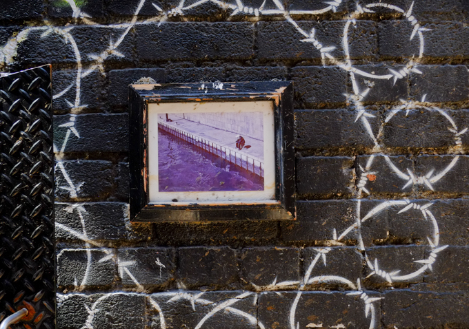 street art in Warburton Lane, Melbourne, a circle of white barbed wire. in the center is a framed photograph of a man fishing beside purple water