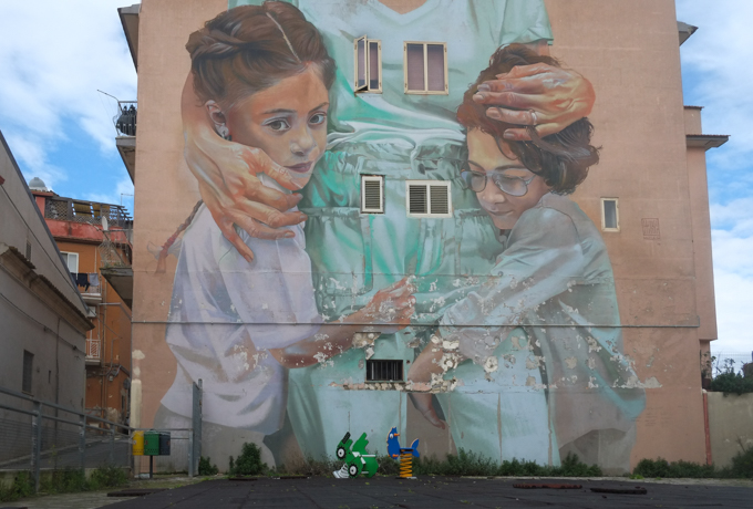 mural by Case Maclaim of two young girls close to their mother who is dressed in turquoise, on the wall of a building beside a playground with a couple of plastic toys 