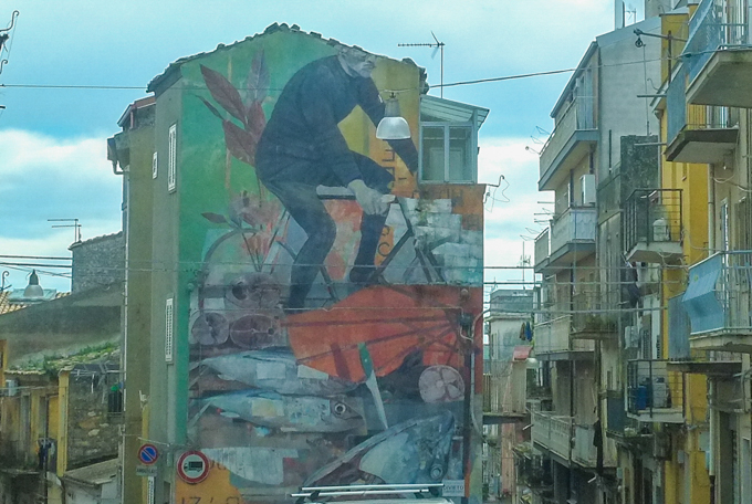 mural by Marak Morik of a man on bicycles, a red umbrella, and a pile of fish. 