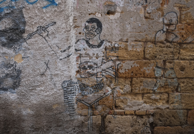 a man, seated, is painting a black face on a stone wall,  faded street art on an old brick and stone wall, textures, alley in Palermo