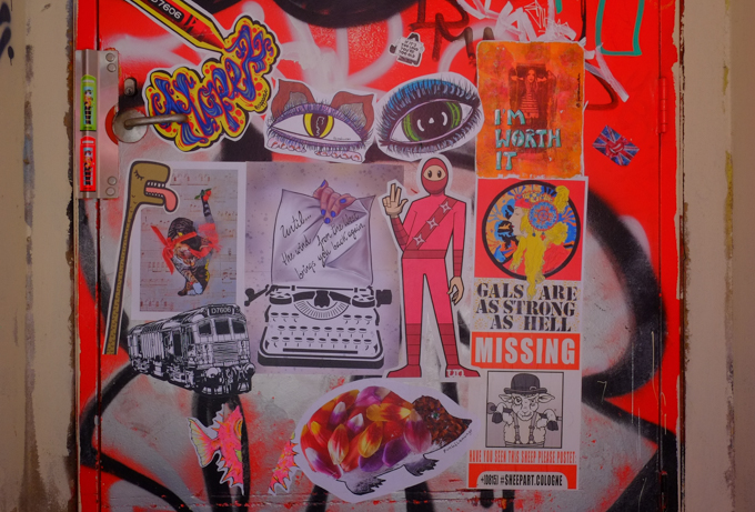 graffiti paste ups and stickers on a wall including catchoo worm with tongue stuck out, a typewriter with a piece of paper with words until the west wind brings you back again. A pair of eyes, a cat collage on sheet music paper, a train by D7606, t bonez from urban ninja squadron dressed in red and giving peace sign with two fingers