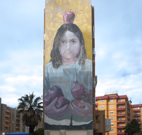 mural by evoca 1, or Elio Mercado of a young woman with an apple on top of her head.  she is holding six other red apples in her hands,