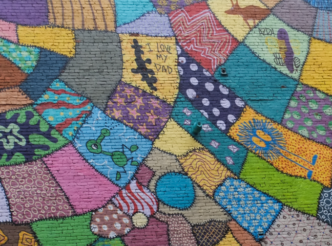 part of mural by Caleb Neelon in Lynn Massachusetts, like patchwork quilt made of many shapes and colours, stitched together with black lines,  with polka dots, squiggles, stars, stripes, 