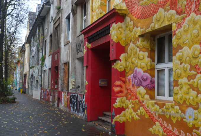 Kiefernstrasse houses, street level, leaves falling on wet sidewalk, one door entrance is bright red, walls have red dragon with gold clouds, 