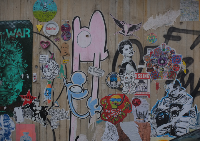 collection of stickers and paper paste ups on a concrete wall in Bonn, one with text that says plus "You're driving me bananas!", a young Sophia Loren, a bird in flight, a green octopus dressed in red and white stripes, and many more