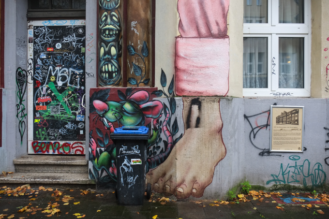 green rat with large pink eras painted on concrete, beside bare foot that is part of a larger mural, rat partially behind a real garbage bin