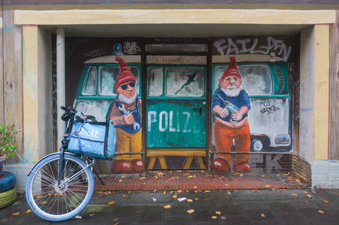 mural of 2 gnomes in red caps and blue tops, working on a green police van.  one gnome is holding a wrench, 