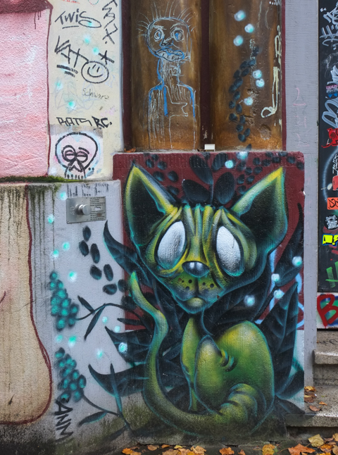 green cat with large white eyes painted on concrete, beside bare foot that is part of a larger mural