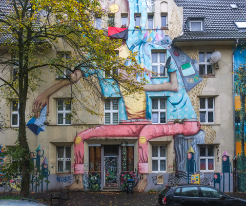 more of mural, blue headscarf, also pink trousers, and bare feet, sitting,  smaller creatures at street level, black outfits, and pyramidal shaped hats