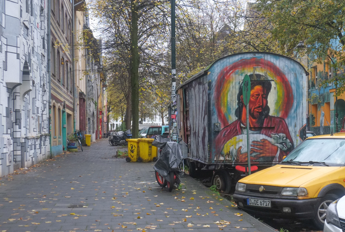 old railway car sits beside sidewalk, covered with street art including an image of Jesus carrying a goat