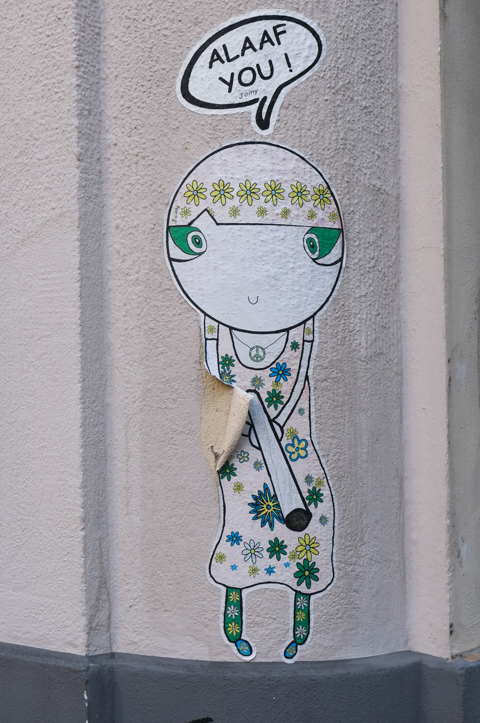 paper paste up by Joiny of a woman saying Alaaf You, wearing a dress wth green and yellow flowers on it, peace sign nacklace