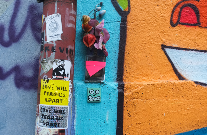 little pieces of graffiti on a wall beside a large mural with orange faces, two stickers that say love will tear us apart, 