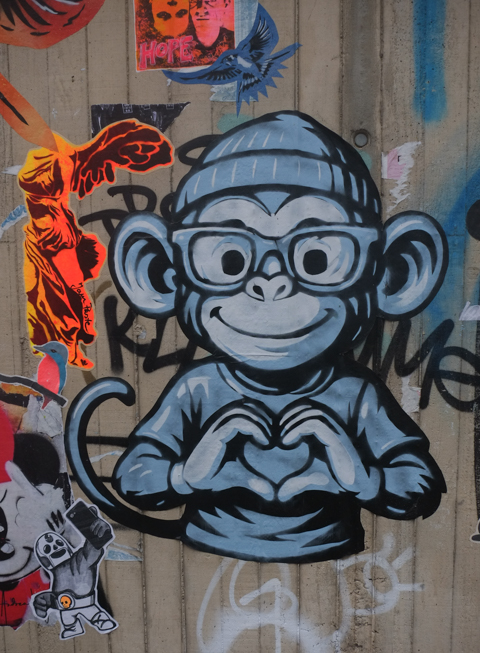 paste ups on a wall, a large blue monkey with glasses and a toque, making a heart sign with his hands, by artist known as Opus, There is also an orange figure by Jam Paste (also graffiti)