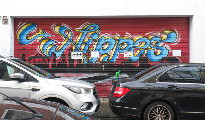 cars parked in front of a mural painted on a metal garage door, red background, the word nippes in calligraphy in blue and yellow by Robin Von Gestern