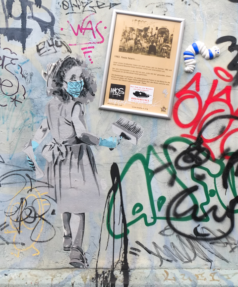 a framed poster describing part of the history of kiefernstrasse beside graffiti stencil of a young girl wearing a covid mask, with a large paint brush in her hand