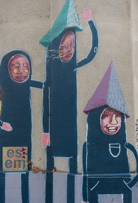 part of larger mural, three smaller creatures    standing at street level, black outfits with just face showing, and pyramidal shaped hats, one hat is turquoise and the other is blue and purple, 
