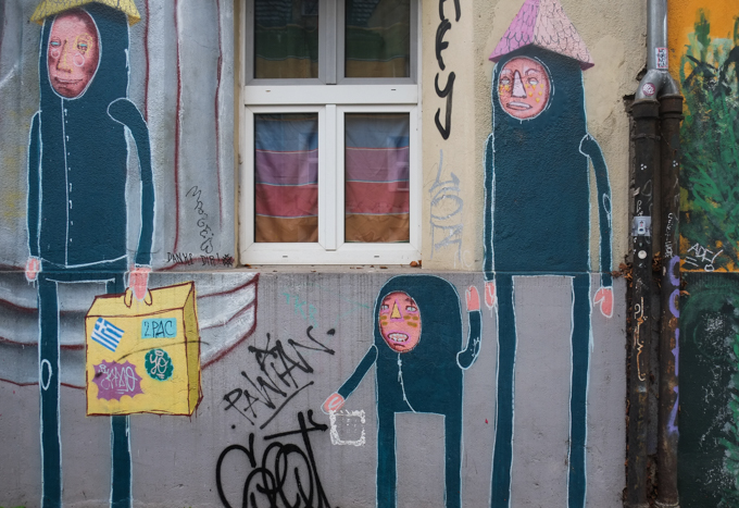 part of larger mural, three smaller creatures    standing at street level, black outfits with just face showing, and pyramidal shaped hats, one hat is yellow and the other is pink.  one of the characters is much shorter than the others and he has no hat