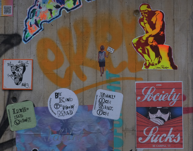 paper paste ups including sign that says Society sucks - be careful, a copy of Rodin's sculpture by Jams Paste, the thinker, a small woman holding a protest sign that says, in German, Frieden Bier and Sonne (peace, beer, sun) by