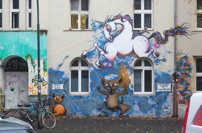part of a mural on kiefernstrasse, white unicorn with laser eyes, and two cats, one sitting in the corner, and one with robot mechanical leg and carrying a flaming sword