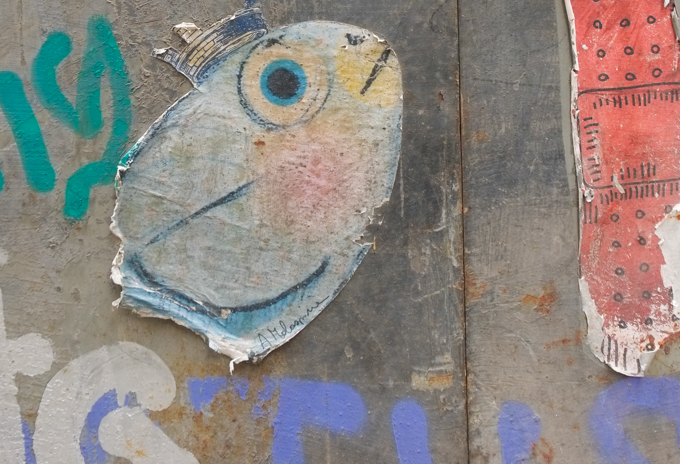 two paper paste ups graffiti on a grey door, one is a little fish with a crown oh its head, and the other is a red band-aid