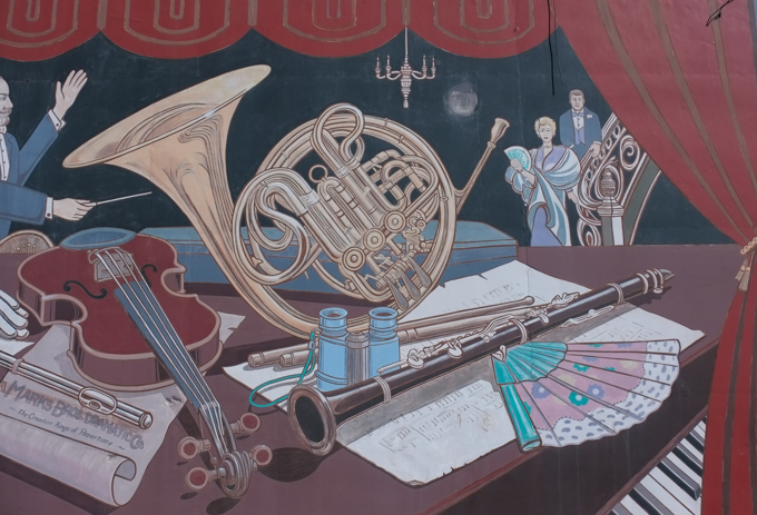 part of borsberry music hall mural in oshawa with images of a french horn, a clarinet, a fan, sheet music,  a violin, and a flute, also a well dressed couple in the background coming down a curved staircase