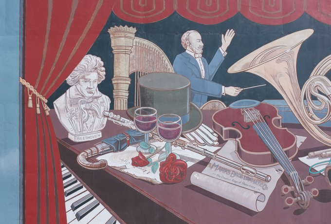 part of borsberry music hall mural in oshawa with images of a flute, bust of Beethoven, harp, ,two glasses of red wine, two red roses, violin, conductor, top hat, piano keys, and stage curtain