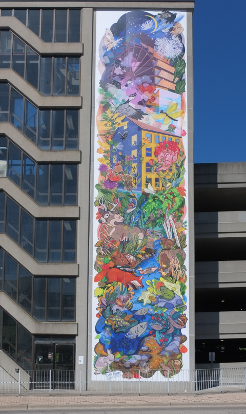 seven storey tall mural on the side of a parking structure, by Dani Crosby, of old grist mill, with large water wheel, colourful, with flowers, fish in the creek, and other wildlife