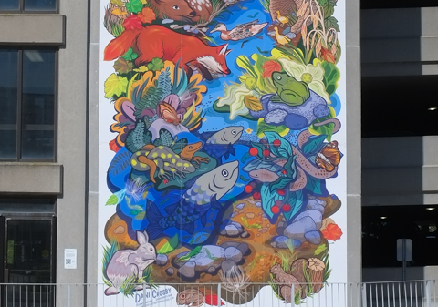 part of seven storey tall mural on the side of a parking structure, by Dani Crosby, of old grist mill, with large water wheel, colourful, with flowers, fish in the creek, and other wildlife, fox, frog, 
