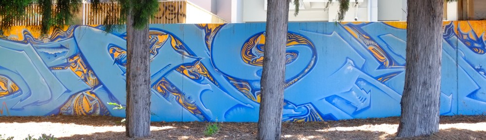 tree trunks in front of a blue mural horizontal, along a low wall or fence in St. Kilda, Melbourne, Australia
