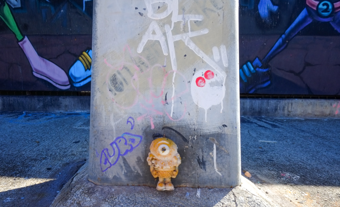 small figure, 3 D, standing at the bottom of a utility pole in front of a mural by East Three