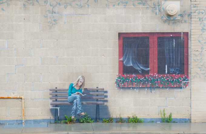mural that looks like life size real image of a woman sitting on a bench beside an open window