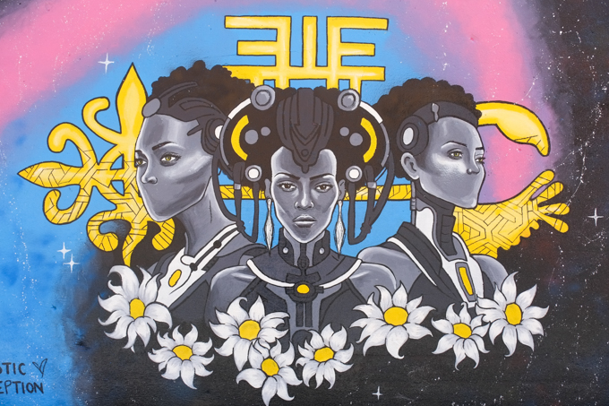 street art mural on wall of railway underpass on Bloor street West by Artistic Perception.  three black women with short curly black hair, clothes in grey shades, with a garland of daisies