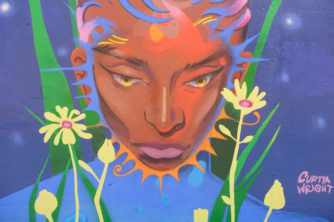 street art mural on wall of railway underpass on Bloor street West by Curtia Wright, portrait of a brown woman with yellowy brown eyes, yellow flowers on either side of her