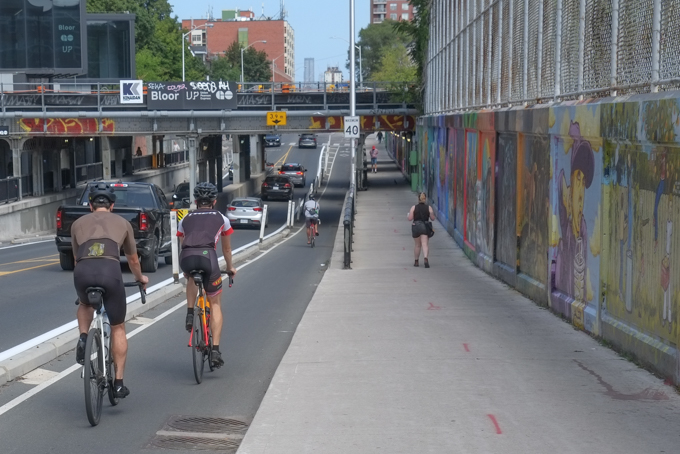 cyclists in bike lane on Bloor West, traveling eastbound and about to go under the railway tracks just west of Perth.  Street art on the walls of the underpass