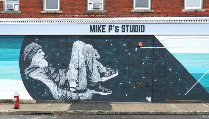 black and white street art mural in Lynn Massachusetts, an older man semi reclining on the ground with his knees up, wearing a hat and running shoes, by Ivan Slazar, on the side of Mike P's Studio