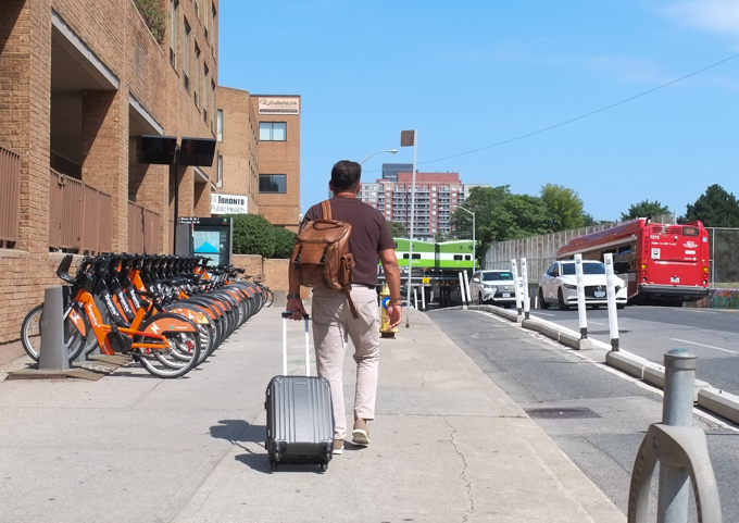 man pulling a suitcase as he walks on the sidewalk on Bloor street, eastbound, with GO train on railway bridge in the distance. 