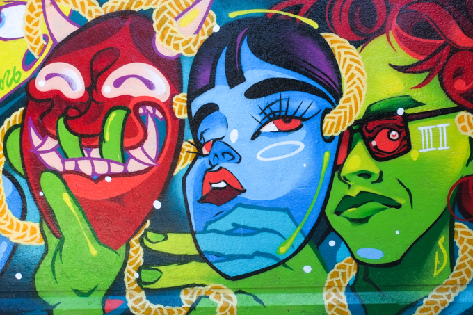 street art mural on wall of railway underpass on Bloor street West by Timmifuk, young man with green face is removing two masks, one is blue and the other is a demon red