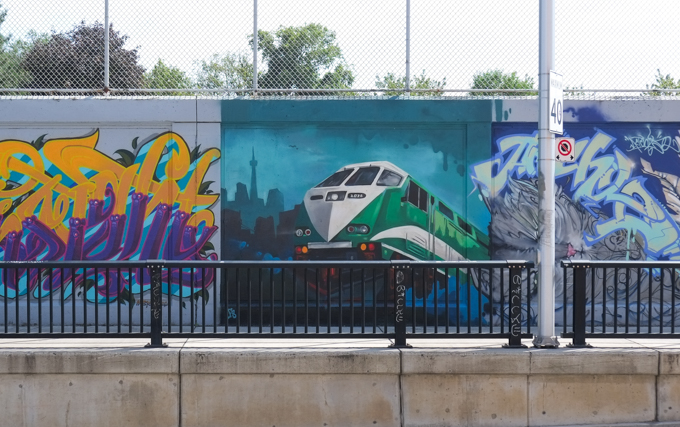 three street art murals on wall of railway underpass on Bloor street West.  an image of a GO rain engine is in the middle