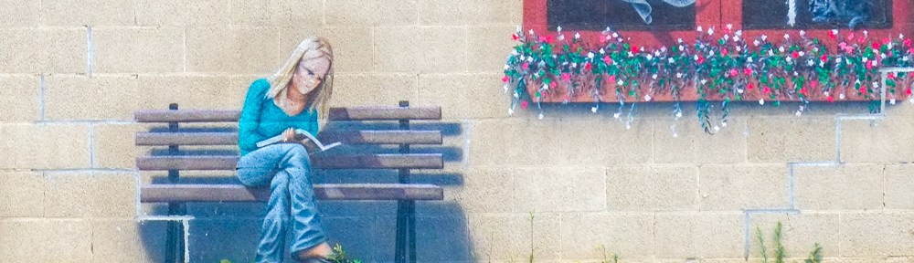 part of a larger mural on a concrete block wall, image of a woman sitting on a bench reading a book, an open window near her with the curtain blowing in the wind