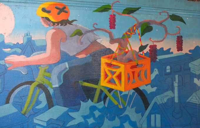 street art mural on wall of railway underpass on Bloor street West by Wenting Li, A cyclist riding a green bicycle past a stylized blue neighborhood skyline, transporting a growing branch with clusters of red fruit in a bright orange milk crate.